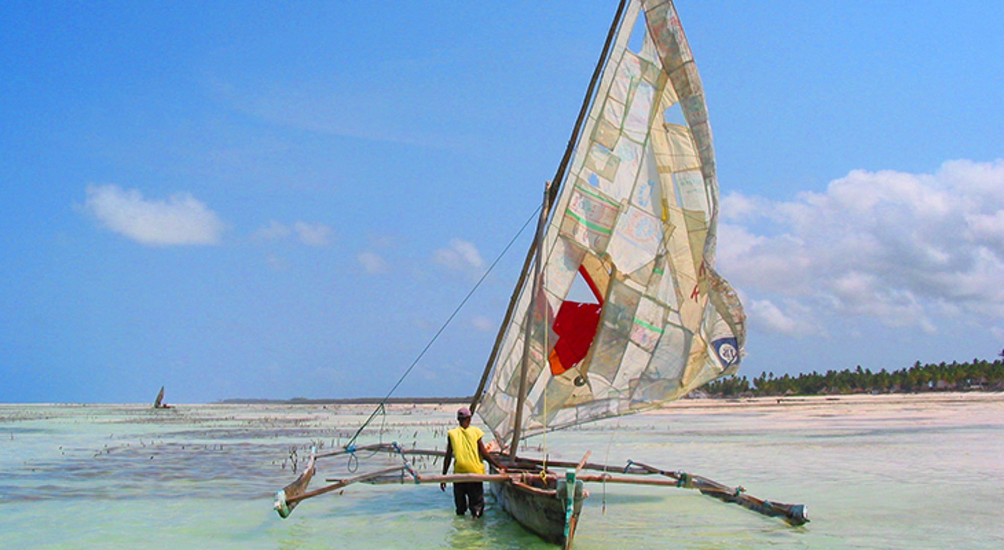 Zeilboot met grote zeilen in ondiep turquoise water bij een tropisch strand onder blauwe lucht.