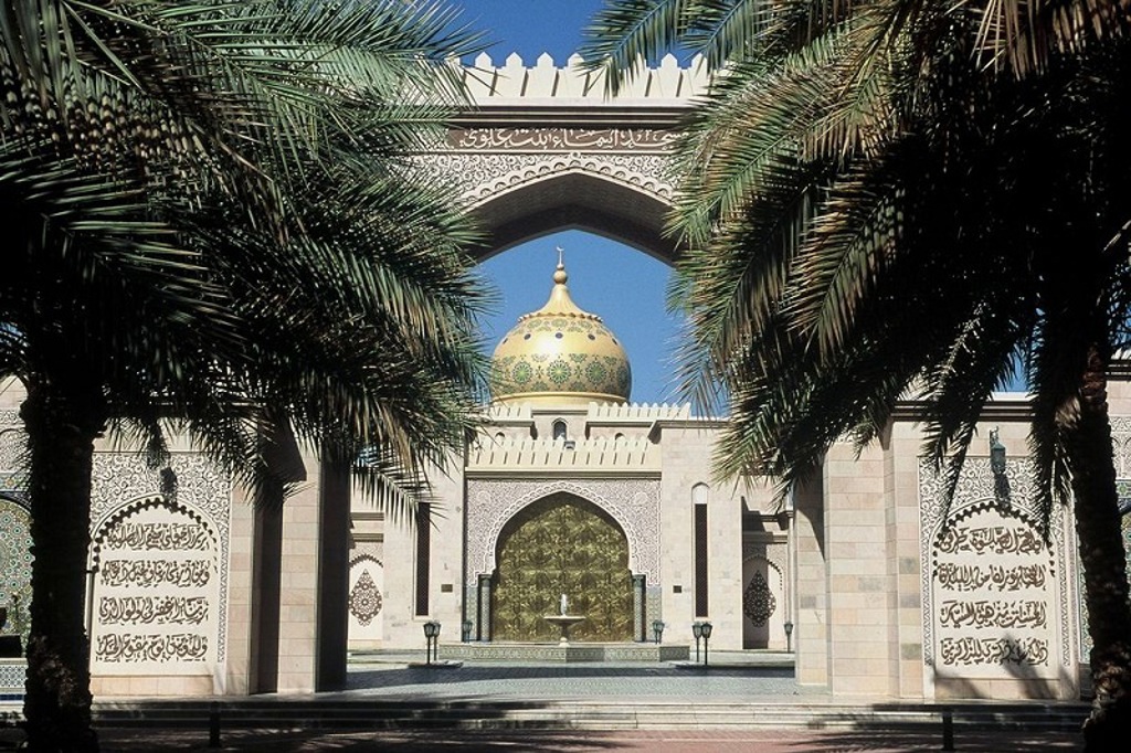 Poort van de Sultan Qaboos Grand Mosque in Muscat, Oman, met gouden koepel tussen palmbomen.