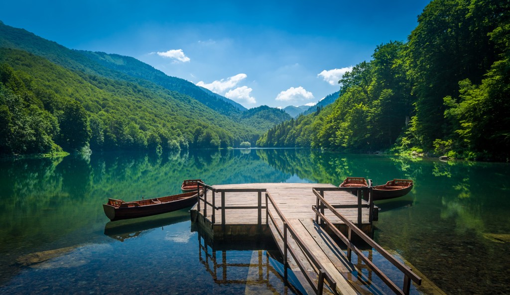 Houten steiger met roeiboten aan een helder bergmeer, omringd door groene heuvels in de Balkan.
