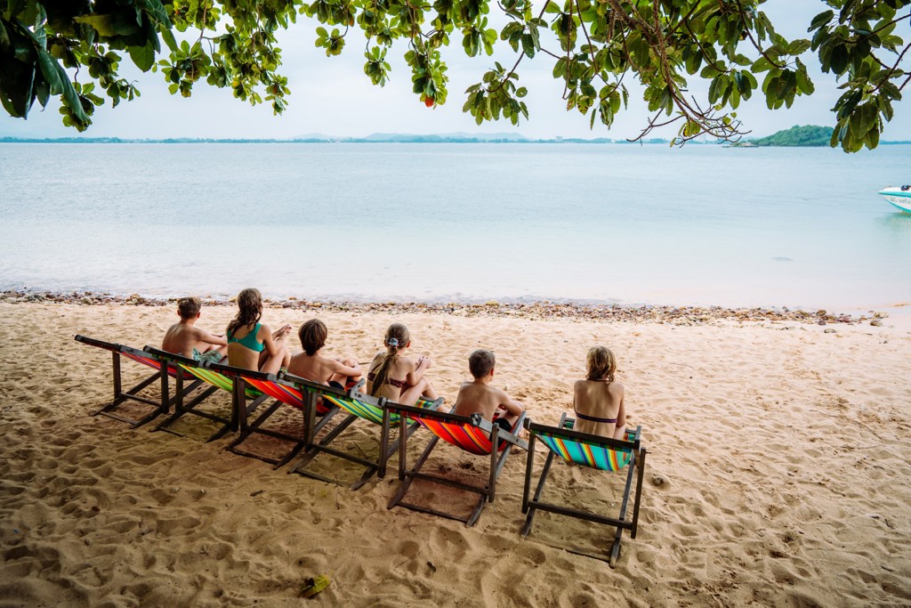 Gezin ontspant op strand in Thailand met ligstoelen onder bomen, uitkijkend over zee.