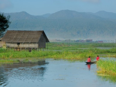 Traditioneel houten huis op palen aan het Inlemeer in Myanmar, met vissersboot en bergen op de achtergrond.