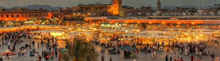 Drukke avondmarkt op het Jemaa el-Fna-plein in Marrakech, met verlichte kraampjes en de Koutoubia-moskee op de achtergrond.