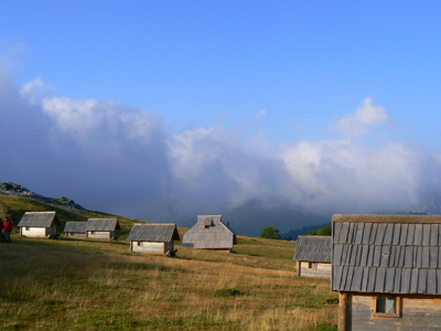 Houten berghutten op een grashelling in Montenegro, met wolken boven de bergen.