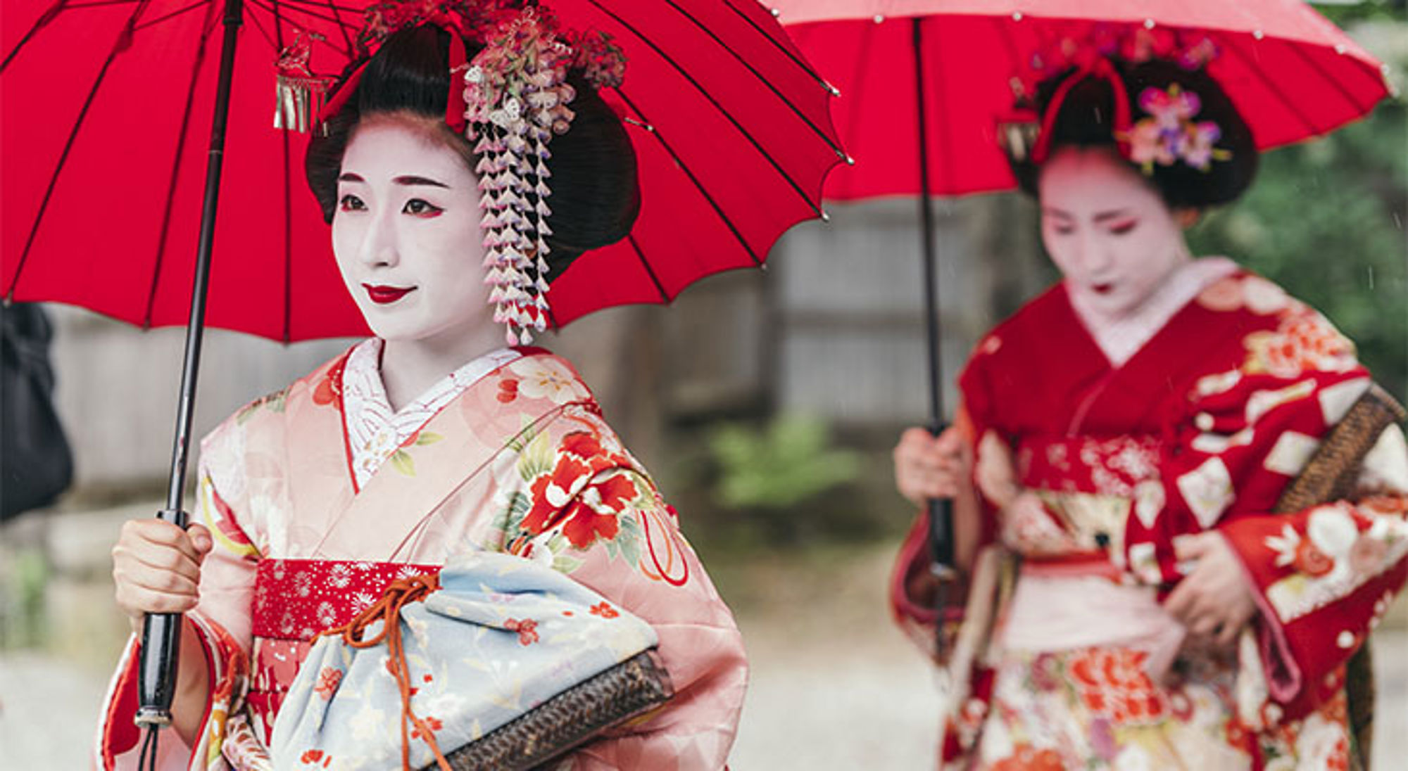 Japanse vrouw in kimono met rode paraplu, met tweede vrouw op de achtergrond.