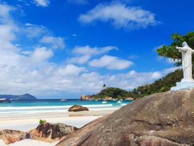 Strand bij Rio de Janeiro met rotsen, bootjes en een Christusbeeld op een rots onder een blauwe lucht.
