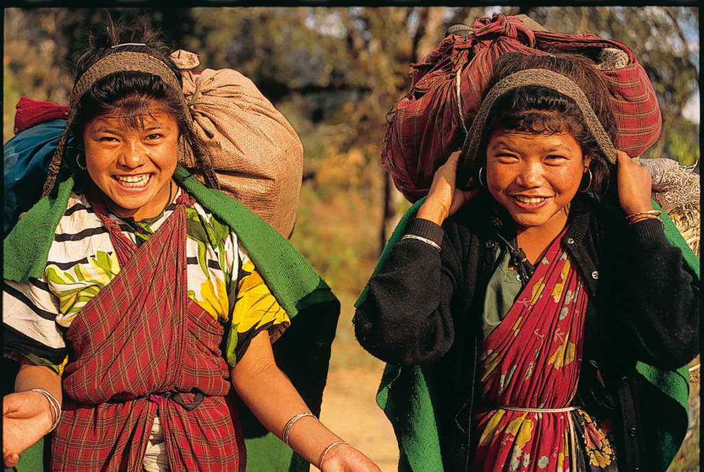 Twee Nepalese meisjes dragen grote bundels op hun rug tijdens een familiereis in Nepal.
