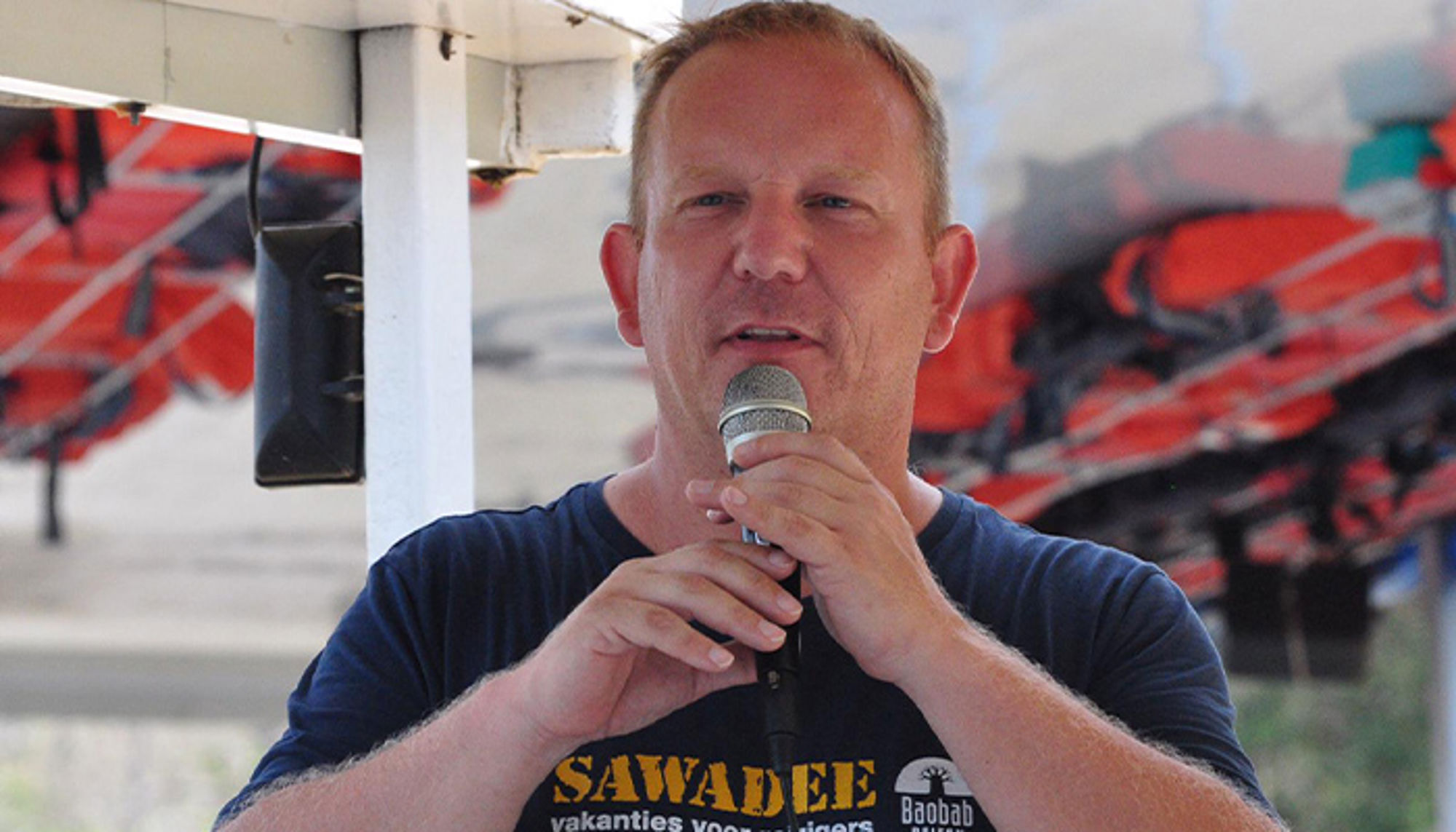Patrick Man spreekt in microfoon tijdens briefing op boot, met reddingsvesten op de achtergrond.