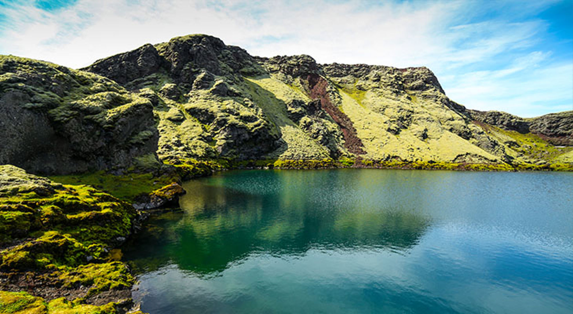 Vulkanisch meer IJsland Groen begroeide rotsen langs een helderblauw bergmeer onder een lichtbewolkte lucht.