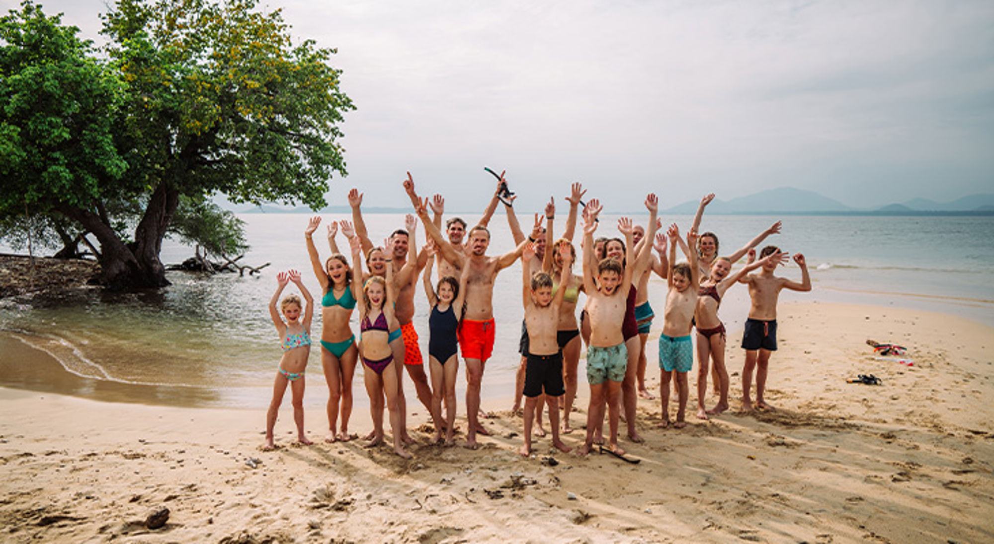 Groep familiereis Groep vakantiegangers juicht op een zandstrand bij zee, met een grote boom op de achtergrond.