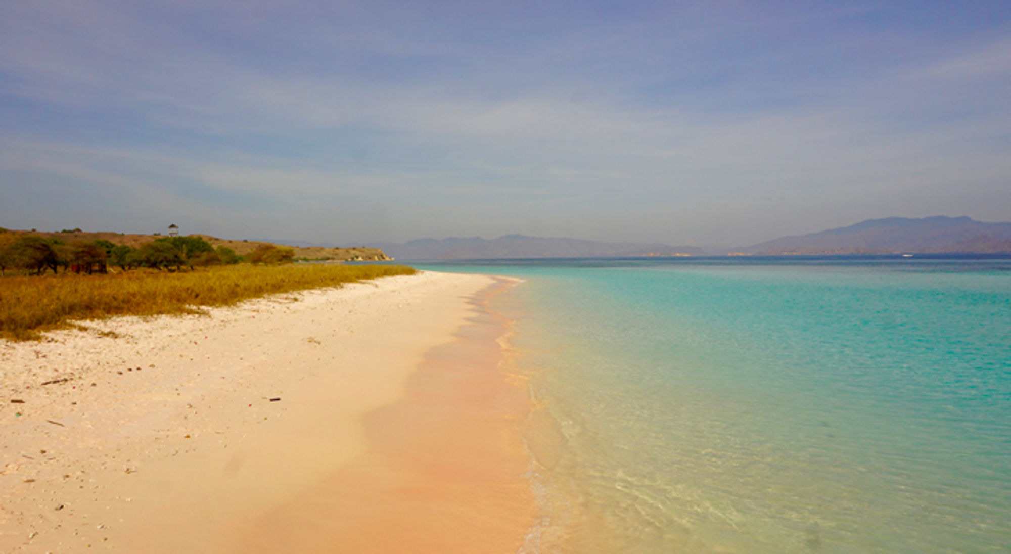Wit zandstrand met helder turquoise water en bergen aan de horizon onder een blauwe lucht.