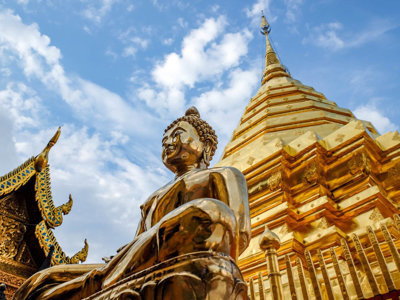 Gouden Boeddhabeeld bij een tempel met gouden chedi in Thailand, onder een blauwe lucht.