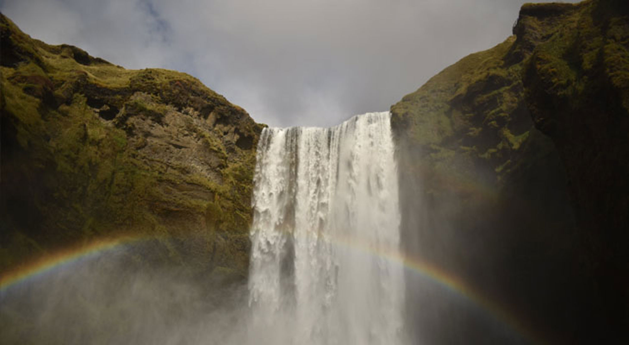 Skógafoss IJsland Waterval Skógafoss in IJsland met regenboog in de nevel, omringd door groene rotswanden.