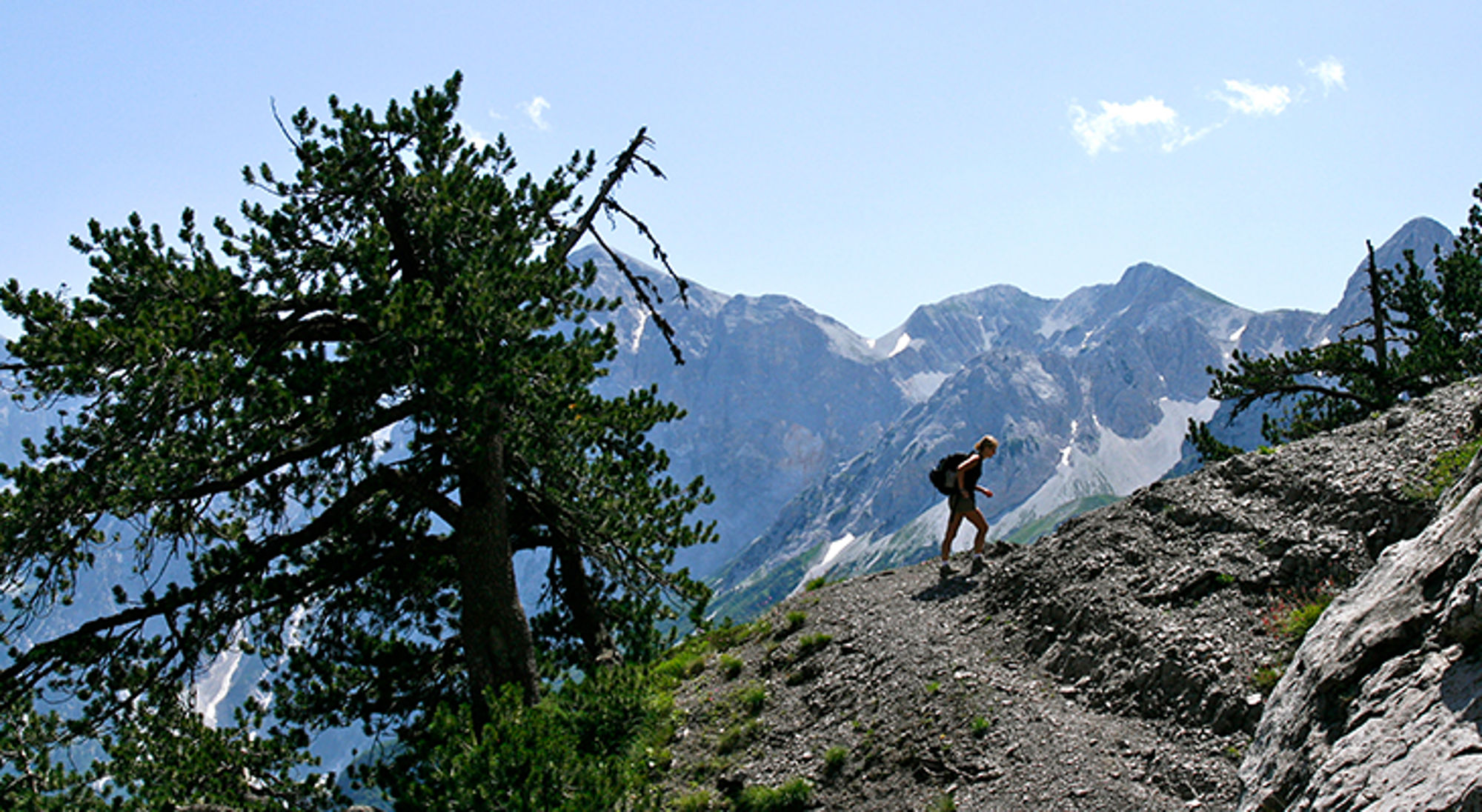 Wandelaar met rugzak op bergpad met uitzicht op ruige bergtoppen en blauwe lucht.