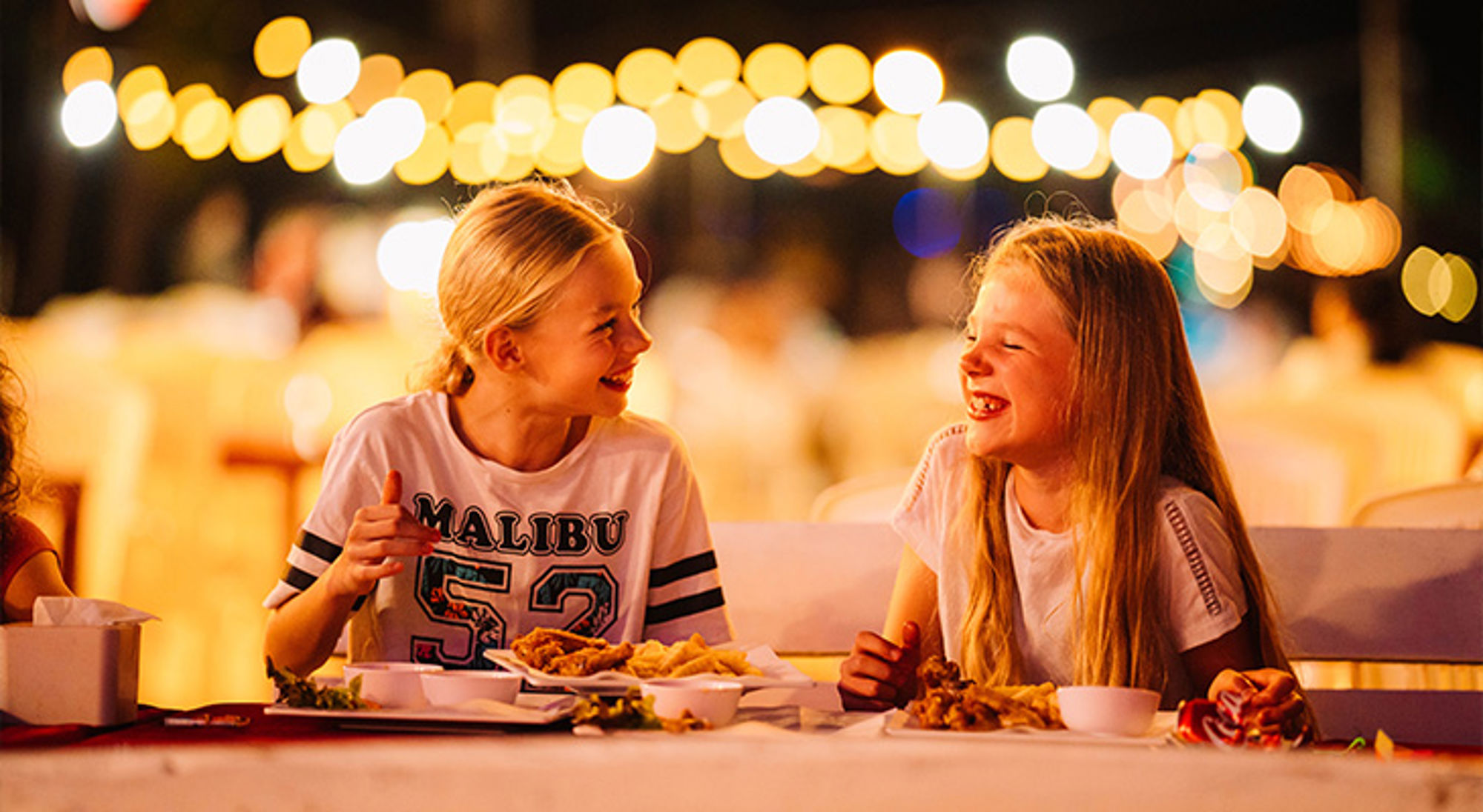 Twee kinderen lachen aan tafel tijdens een avondmaaltijd met sfeervolle lichtjes op de achtergrond.