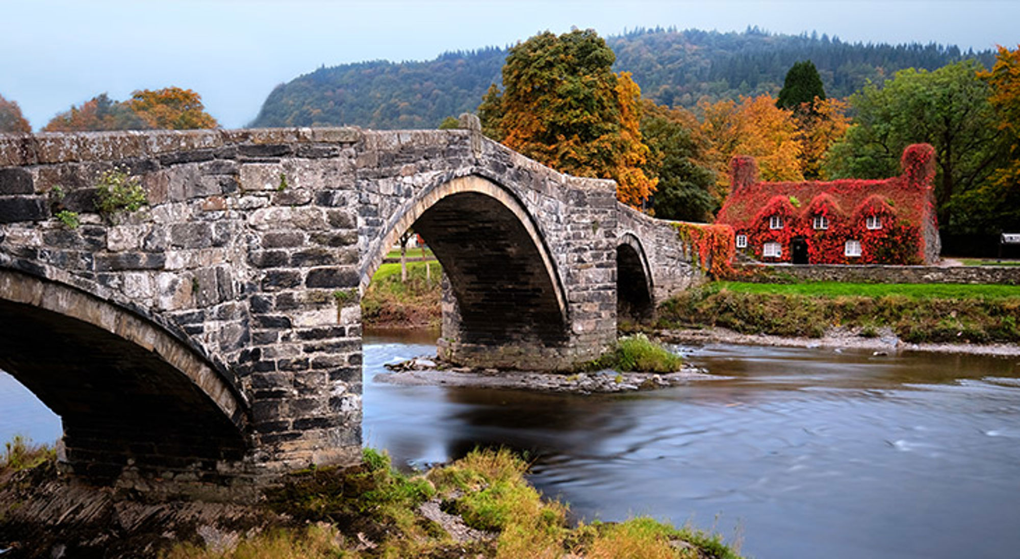 Snowdonia National Park, Wales Stenen boogbrug over een rivier met herfstkleuren en een rood begroeid huis op de achtergrond.