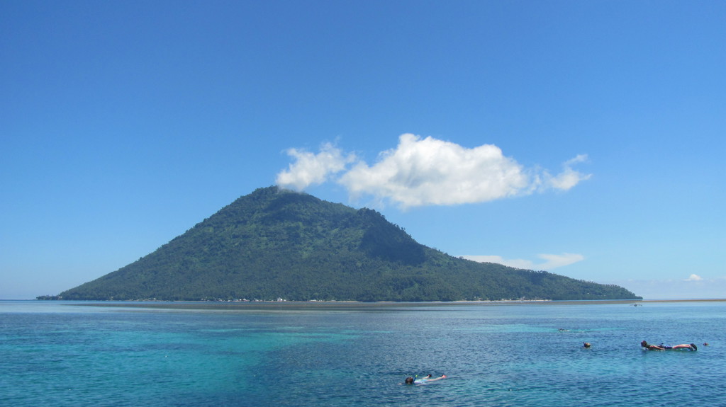 Groene vulkanische eilandberg in zee bij Sulawesi, met snorkelaars in helderblauw water.