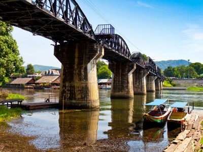 Brug over de rivier Kwai bij Kanchanaburi, Thailand, met longtailboten aan de oever.