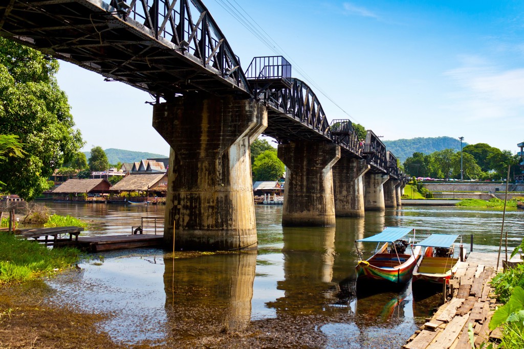 Brug over de rivier Kwai bij Kanchanaburi, Thailand, met longtailboten aan de oever.