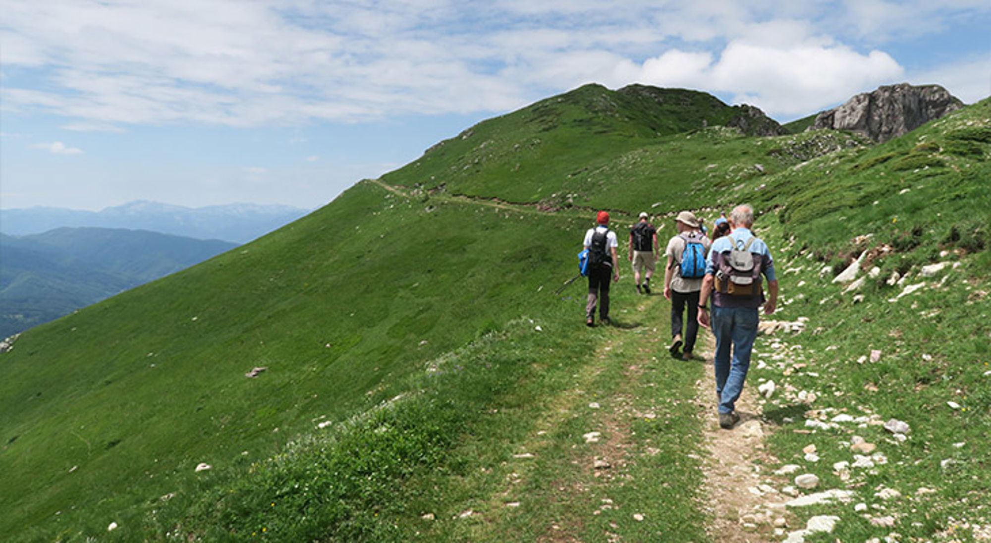 Wandelreis Montenegro Wandelaars met rugzakken op een bergpad langs groene heuvels onder een blauwe lucht.