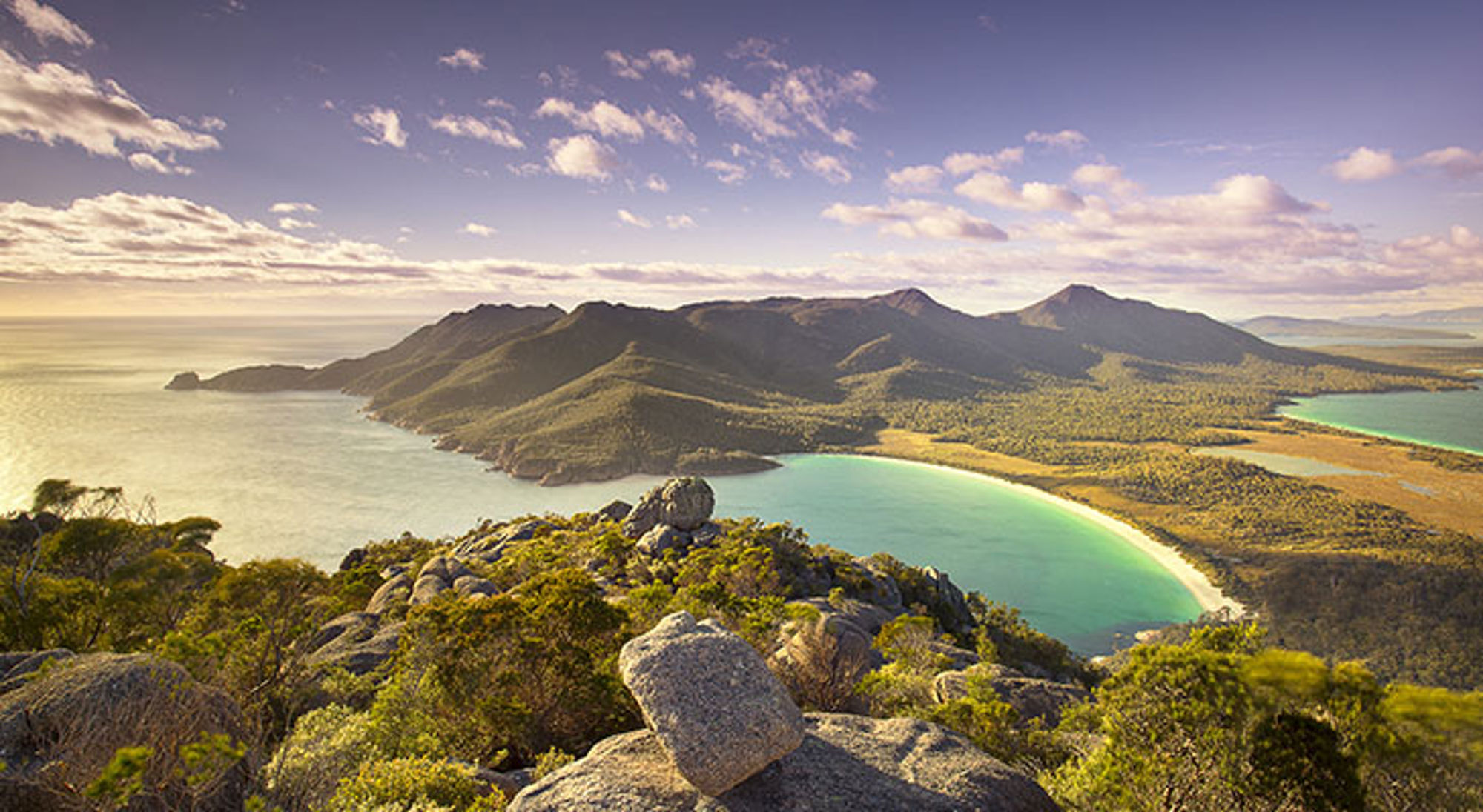 Panoramisch uitzicht op een bergachtig schiereiland met turquoise baai en zandstrand onder een blauwe lucht.