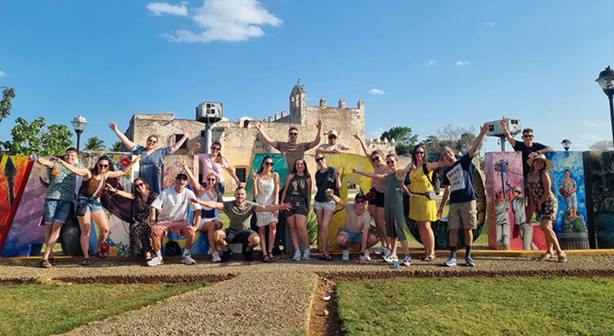 Groep reizigers poseert voor kleurrijke kunstpanelen bij een historisch fort onder een blauwe lucht.