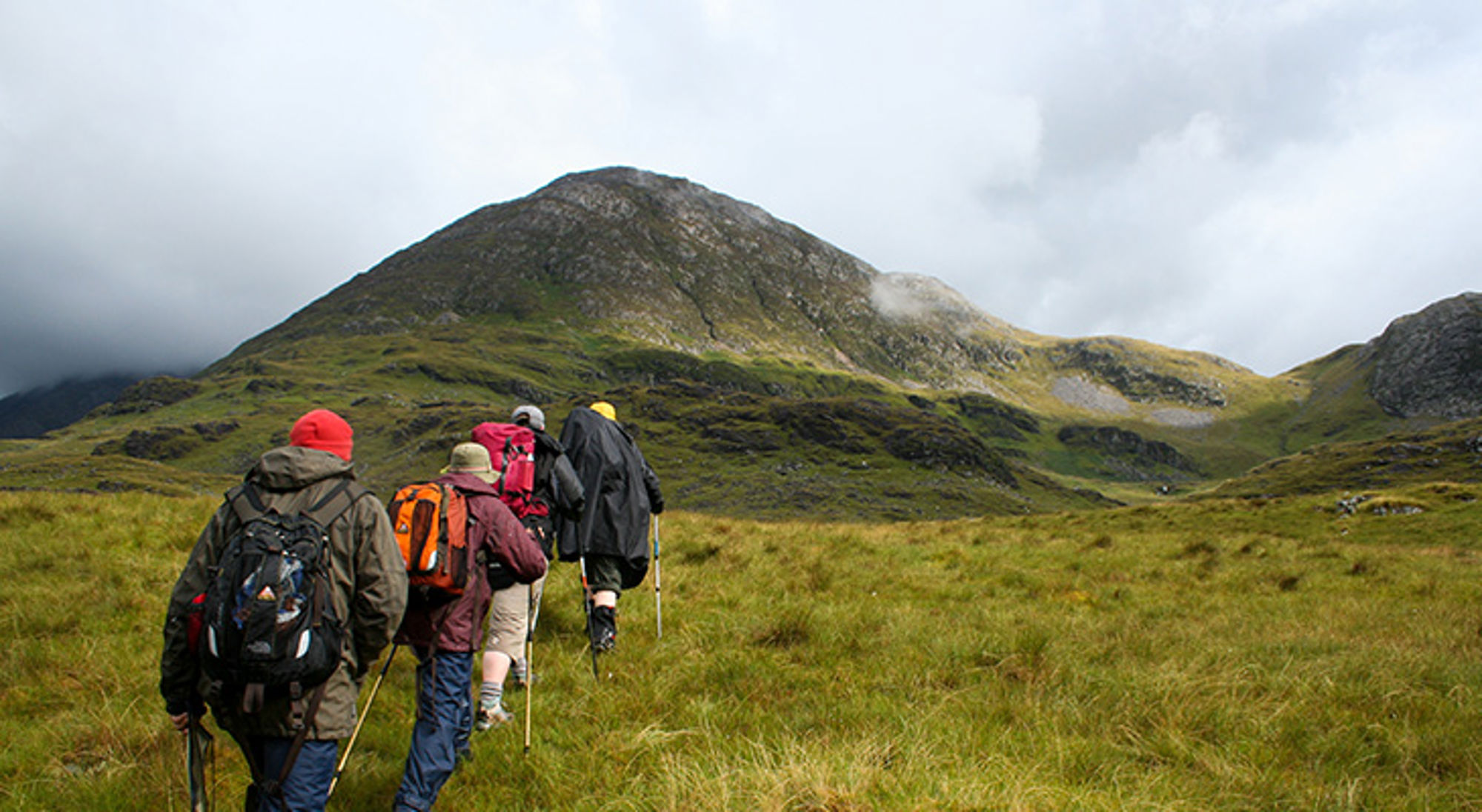 Connemara National Park, Ierland Wandelaars met rugzakken trekken door een groen berglandschap richting een hoge bergtop onder bewolkte lucht.