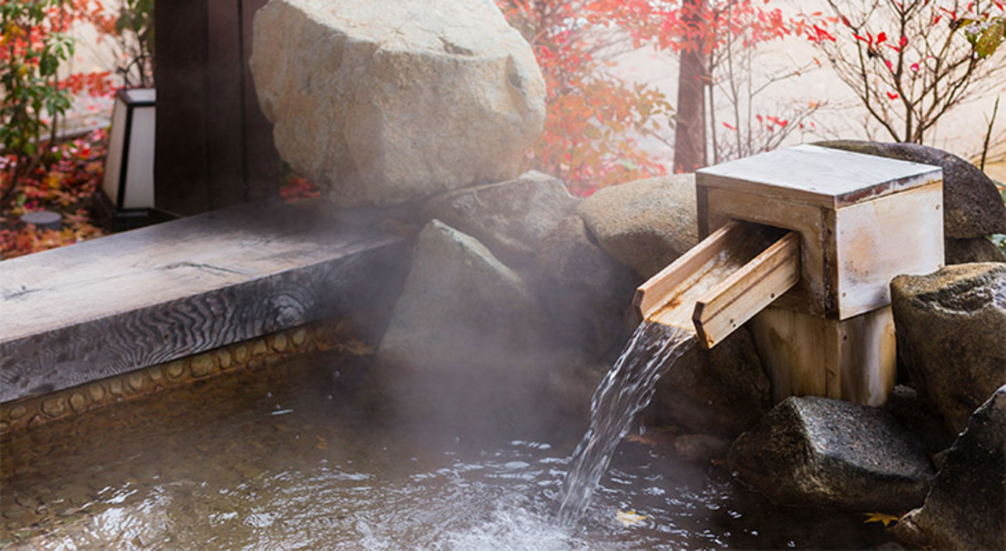 Onsen Stomende Japanse onsen met houten watergoot en rotsen, omringd door herfstbladeren.