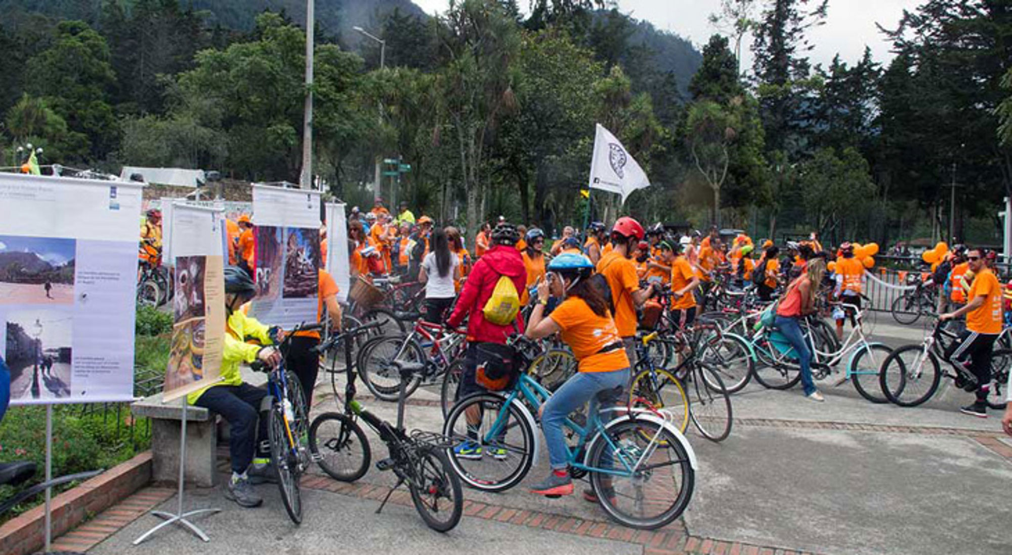 Groep fietsers in oranje shirts verzamelt zich bij een fietsevenement in het park, met informatieborden op de voorgrond.