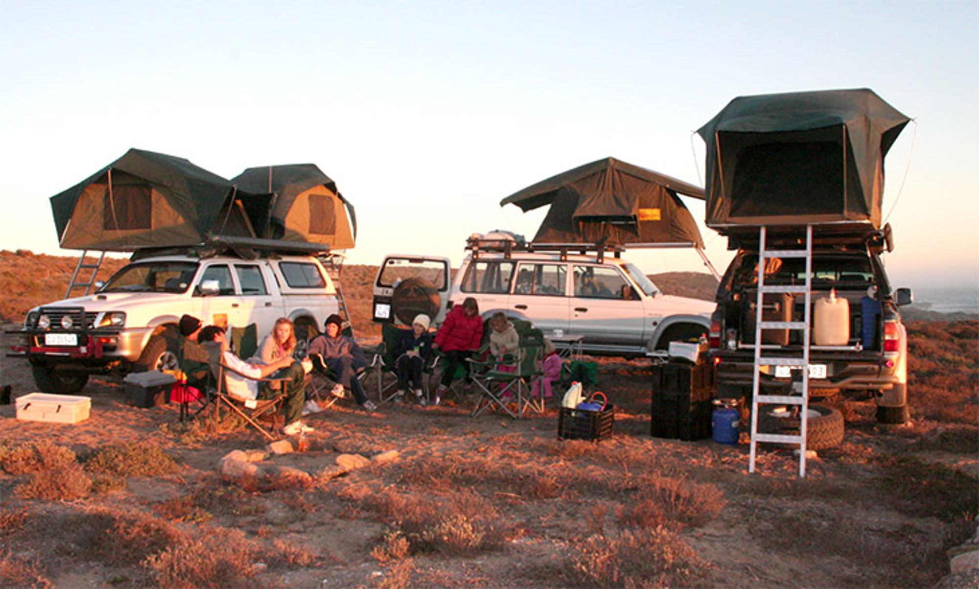 Kamperen in Zuidelijk Afrika Kampeerplek met drie 4x4-auto’s met daktenten en reizigers rond een kampvuur bij zonsondergang.