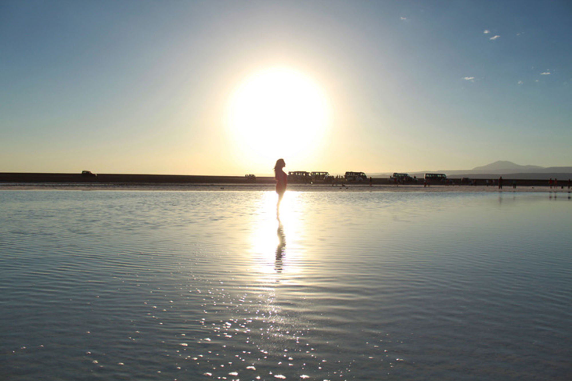 Atacama-woestijn, Chili Silhouet van een persoon in ondiep water bij zonsondergang, met jeeps op de achtergrond.
