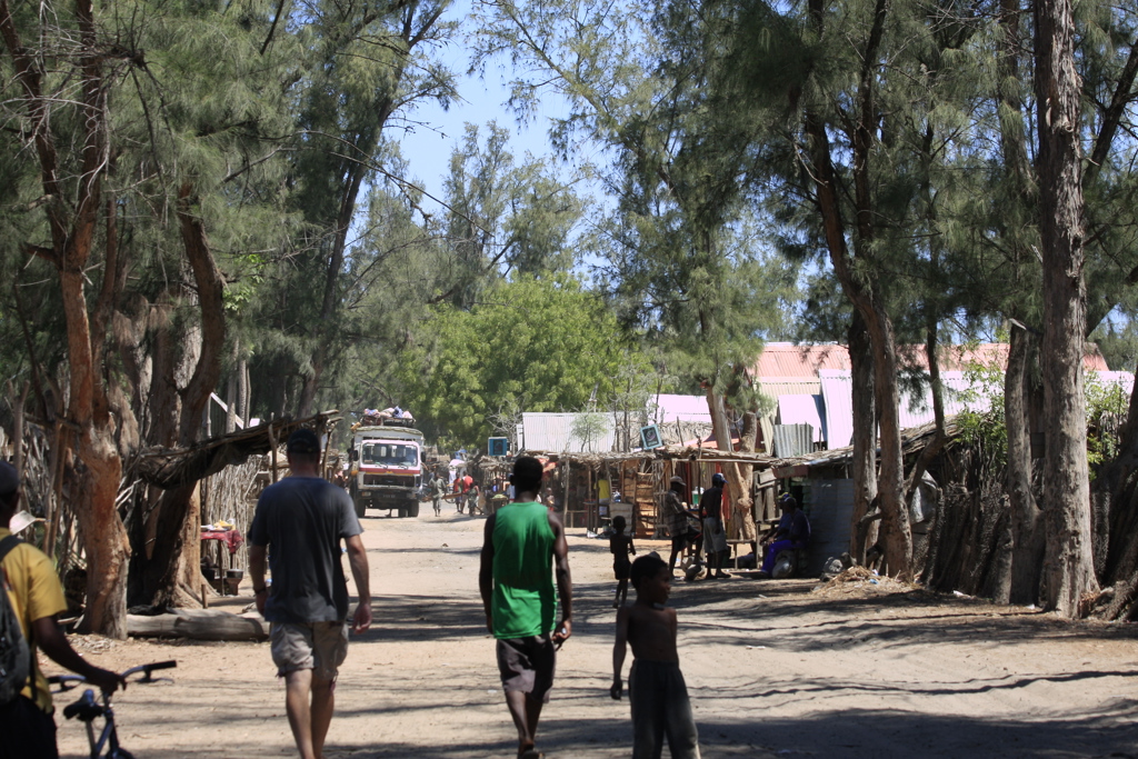 Straatbeeld in Madagascar met lokale marktstalletjes langs een zandweg en mensen die wandelen onder bomen.