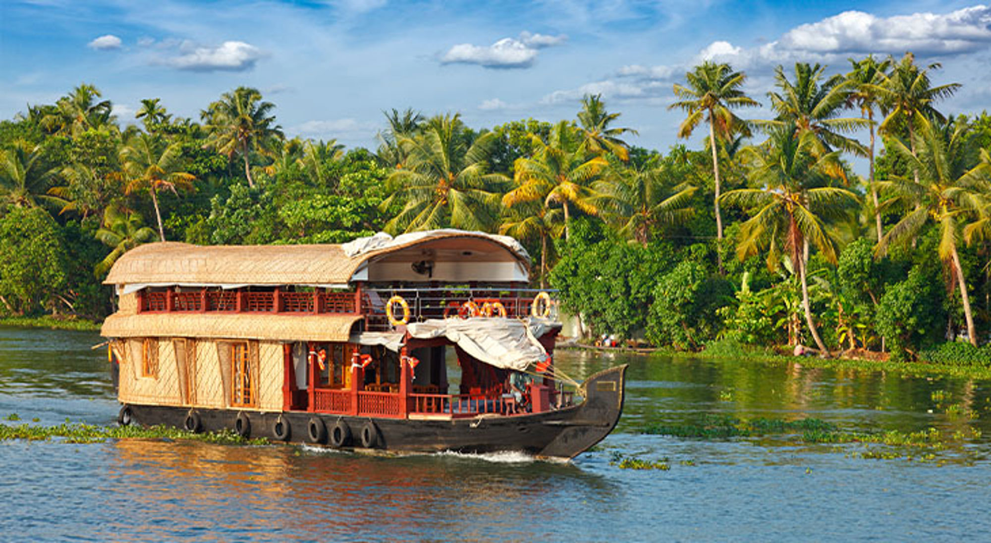 Houseboat Kerala India Traditionele woonboot vaart over een rivier langs een groene oever met palmbomen en tropische begroeiing.