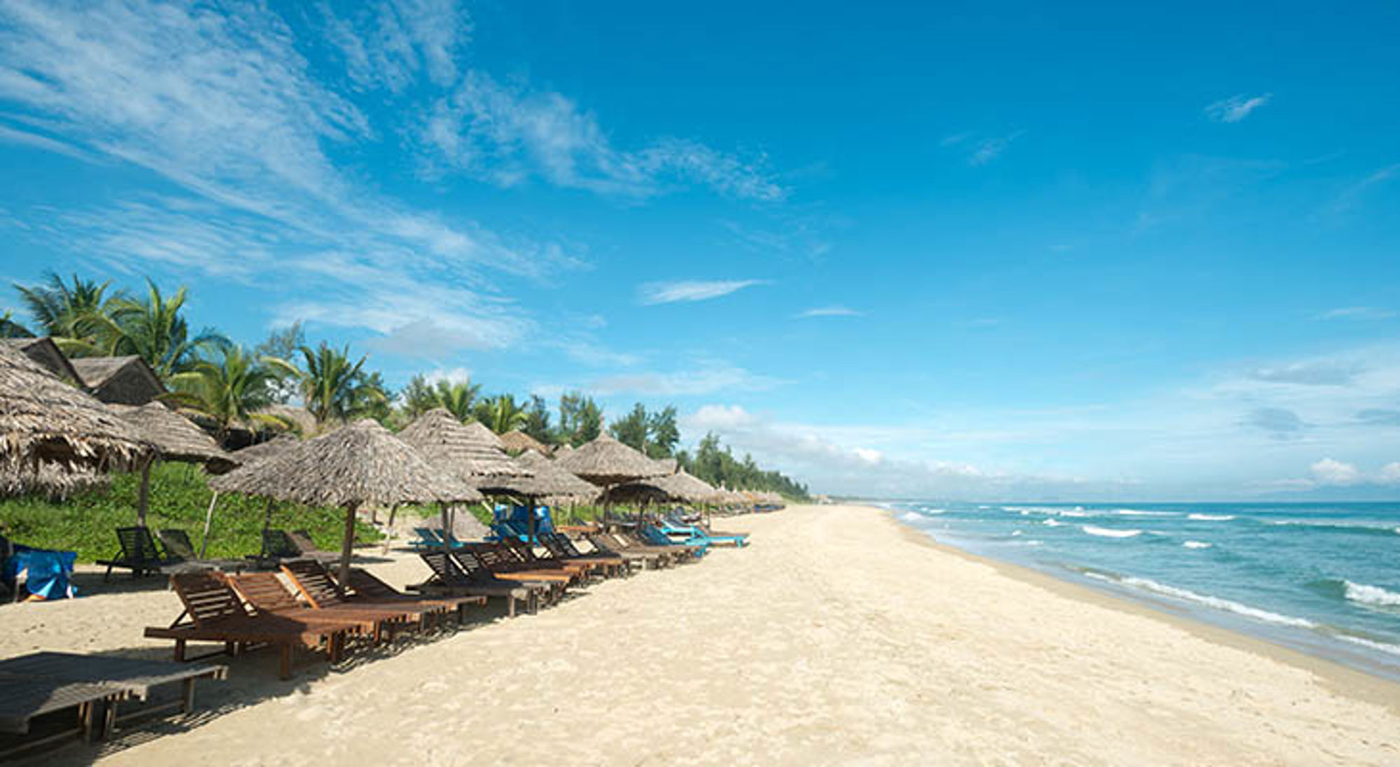 Strand in Vietnam met rieten parasols en ligbedden langs de kust onder een blauwe lucht.