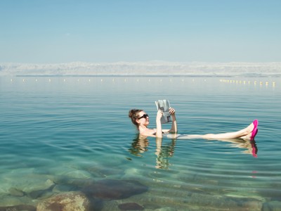 Vrouw drijft in de Dode Zee en leest een krant, met bergen op de achtergrond.