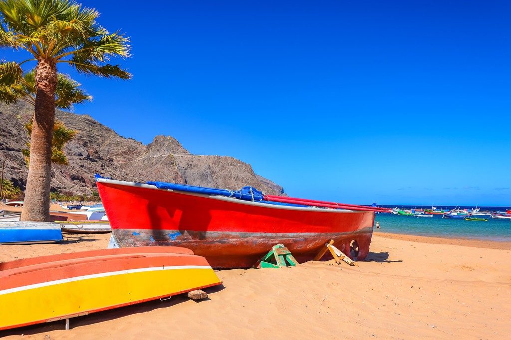 Rode vissersboot op het strand van Tenerife met palmbomen, bergen en blauwe zee op de achtergrond.