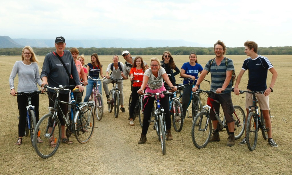 Groep fietsers tijdens familiereis in Tanzania, met savanne en bergen op de achtergrond.