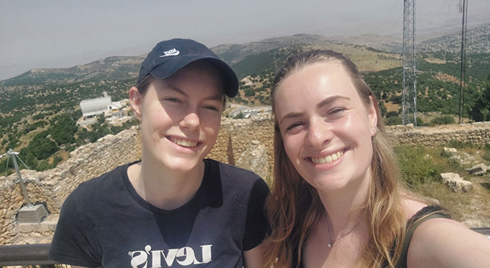 Twee reizigers maken een selfie bij oude stenen ruïnes met heuvelachtig landschap op de achtergrond.