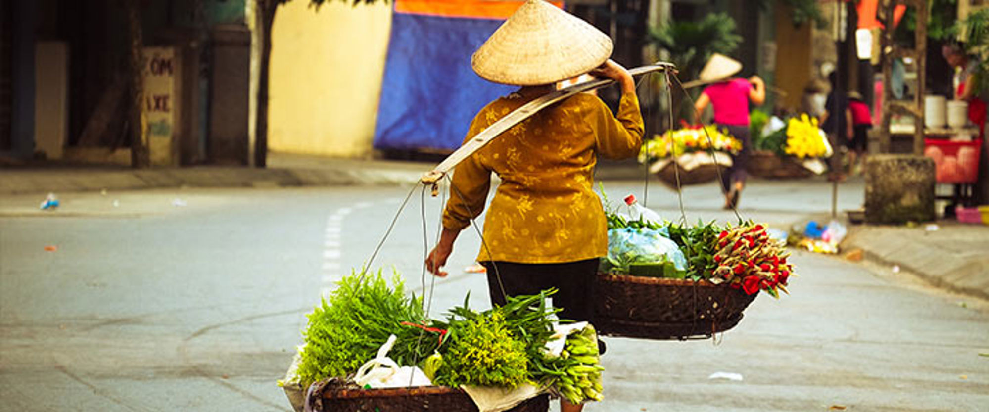 Verkoper in Vietnam Straatverkoper met draagjuk en manden vol groenten en bloemen op een drukke straat in Vietnam.