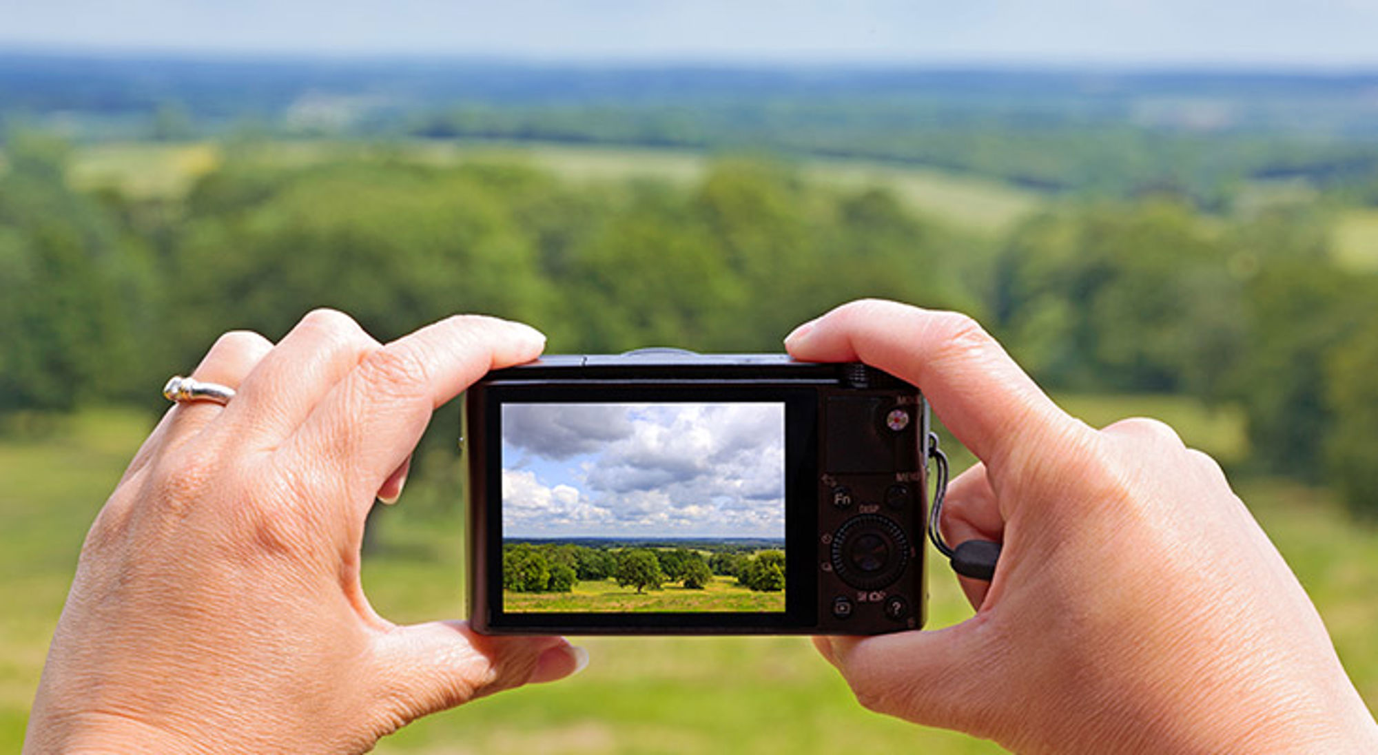 Handen maken een foto van een groen landschap met bomen en wolkenlucht.