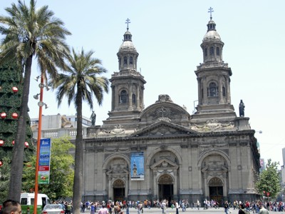 Gevel van de Catedral Metropolitana in Santiago de Chile, met palmbomen en mensen op het plein.
