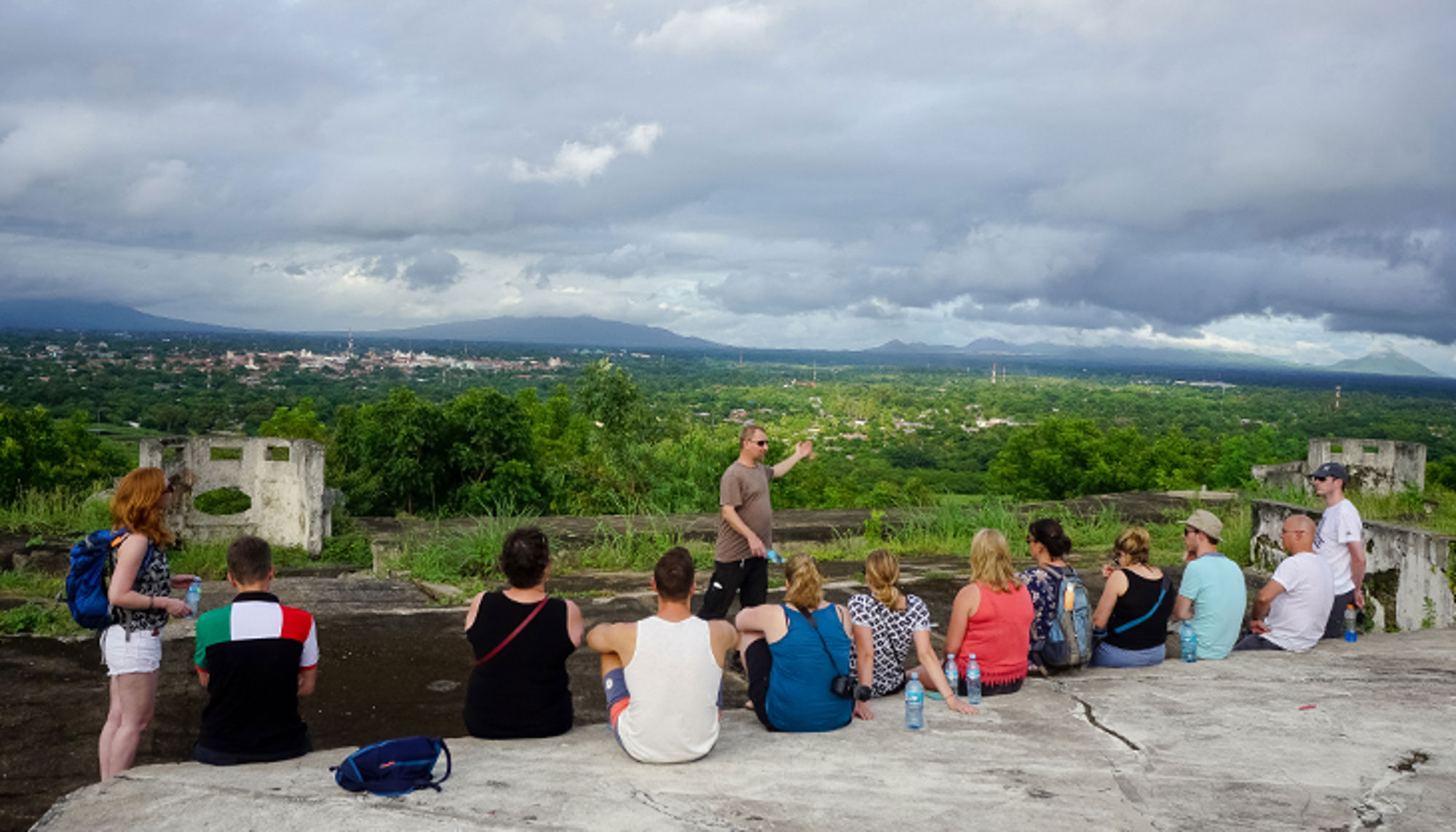 Groep reizigers luistert naar gids bij uitzichtpunt met panorama over groene vallei en bergen onder bewolkte lucht.