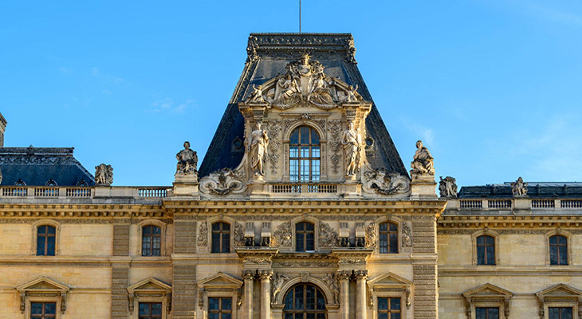 Louvre, Parijs Detail van de gevel en het dak van het Louvre in Parijs, met beeldhouwwerken tegen een blauwe lucht.