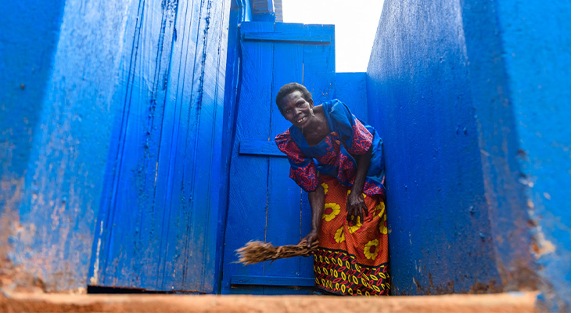 Donatie wc-huisjes Uganda Vrouw in kleurrijke kleding veegt met een bezem bij een felblauwe poort.