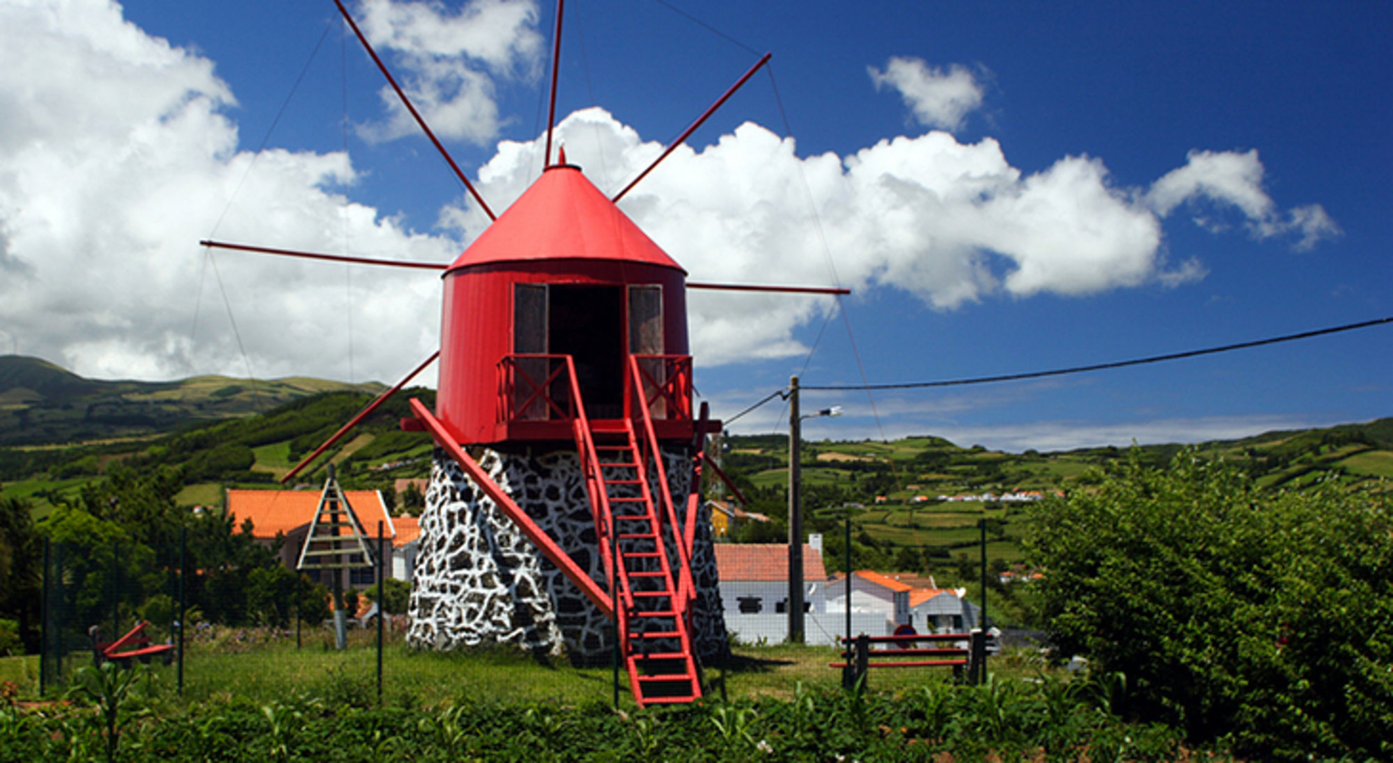 Rode windmolen met stenen basis in groen landschap onder blauwe lucht met wolken.