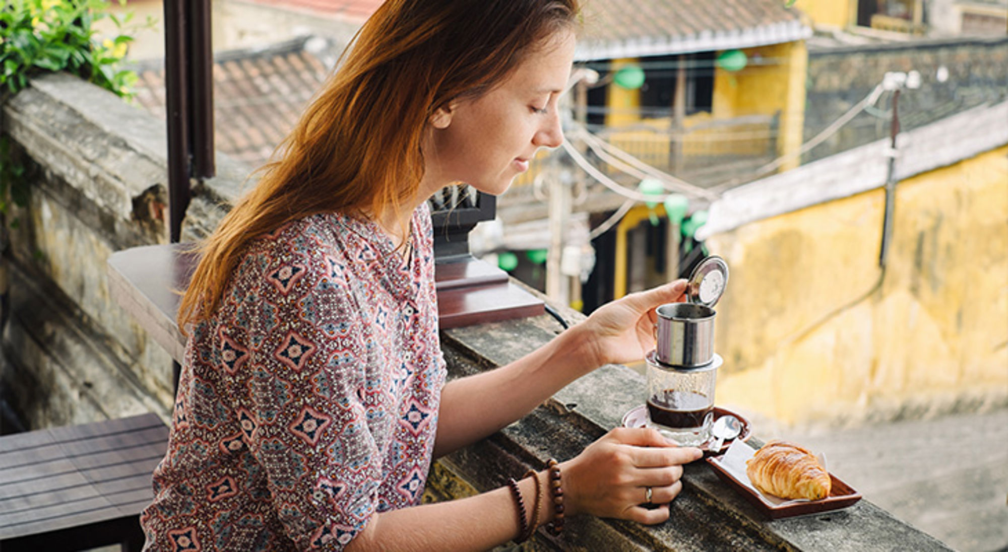 Koffie café Vietnam Vrouw drinkt Vietnamese koffie met phin-filter op een balkon, met een croissant op een bordje.
