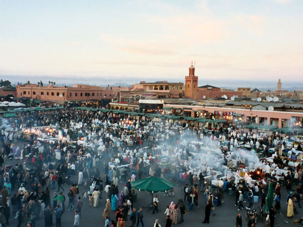 Drukte op het Djemaa el Fna-plein in Marrakech met eetkraampjes en rook van grills tijdens zonsondergang.