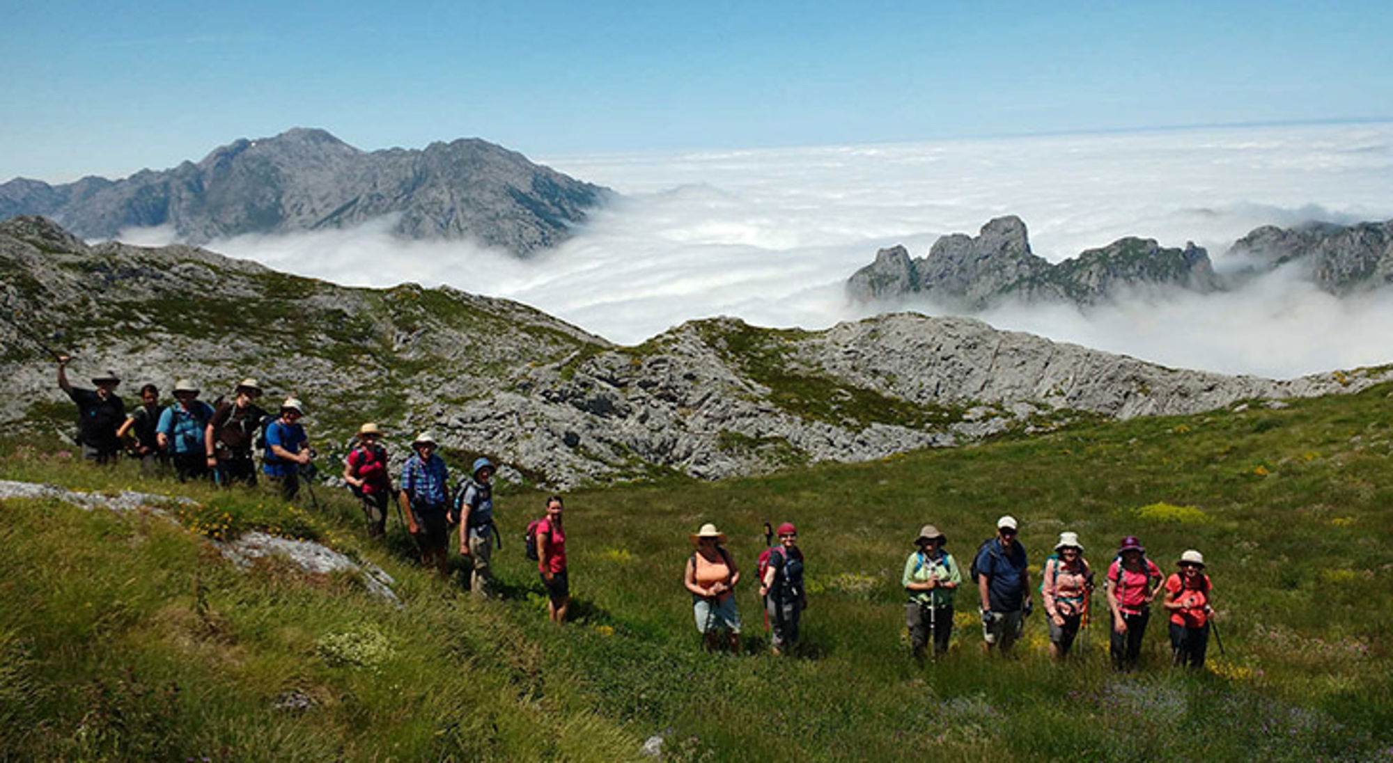 Wandelvakantie Spanje Wandelaars op bergpad door groene alpenweide met rotsen en wolkenzee op de achtergrond.
