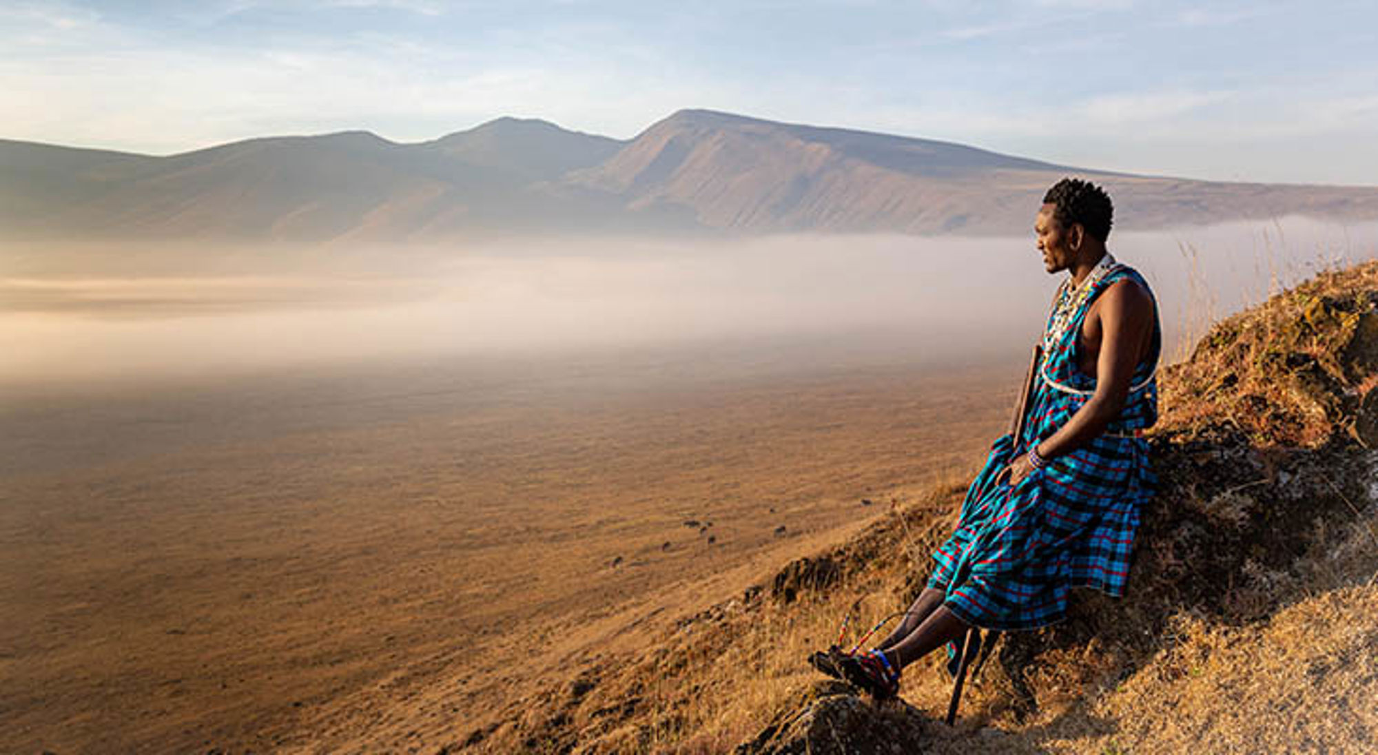 Jonge Masai krijger zittend in de krater van Ngorongoro park kijkend naar zonsopgang Man in traditionele Maasai-kleding kijkt uit over de Ngorongoro-krater in Tanzania.