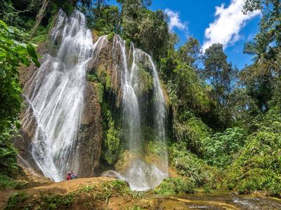 Waterval in de Cubaanse jungle, omringd door tropisch groen en rotsen onder een blauwe lucht.