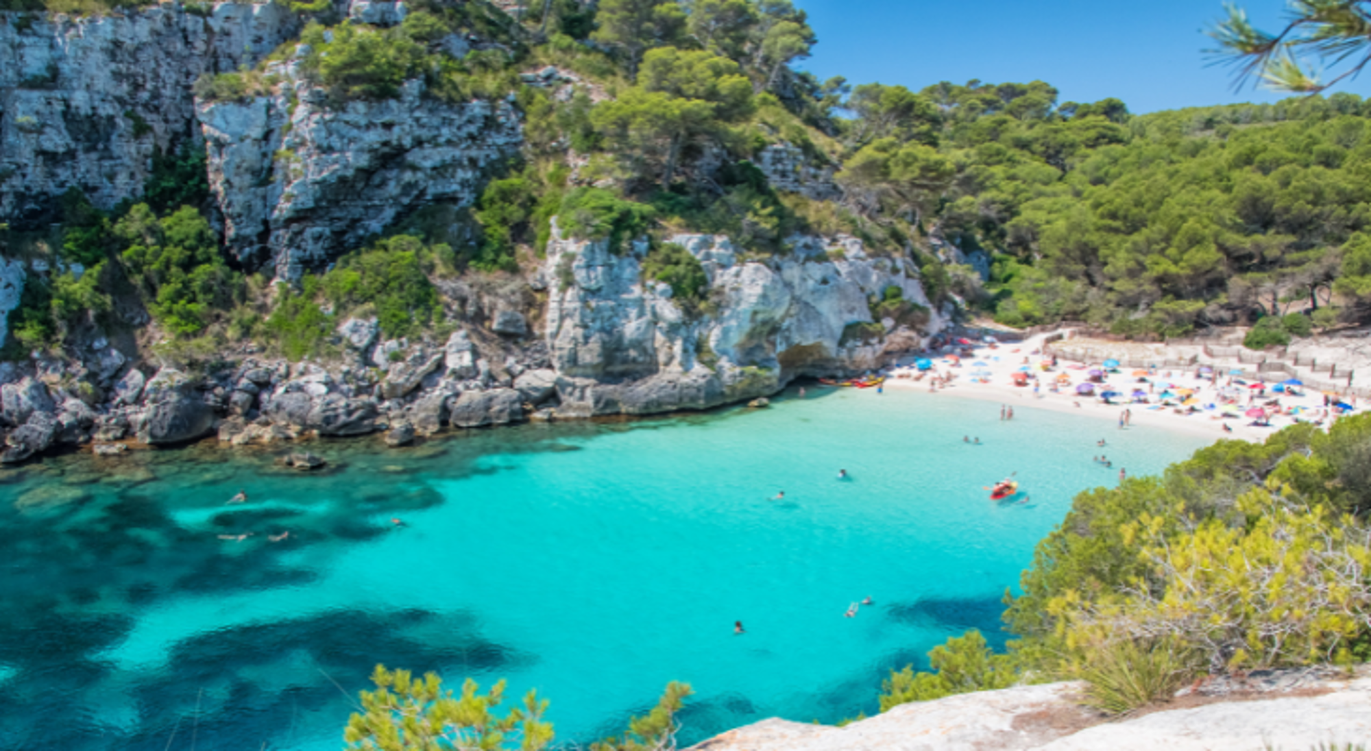 Baai met turquoise zeewater, zandstrand en rotsachtige kliffen met pijnbomen.